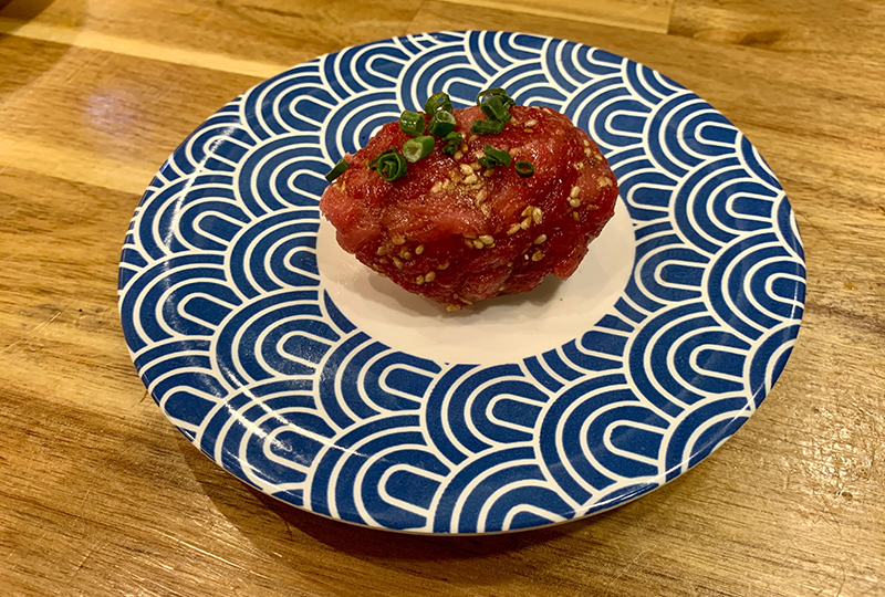 和風藍色盤子上精緻擺放的生牛肉壽司，撒有芝麻和蔥綠色調料。