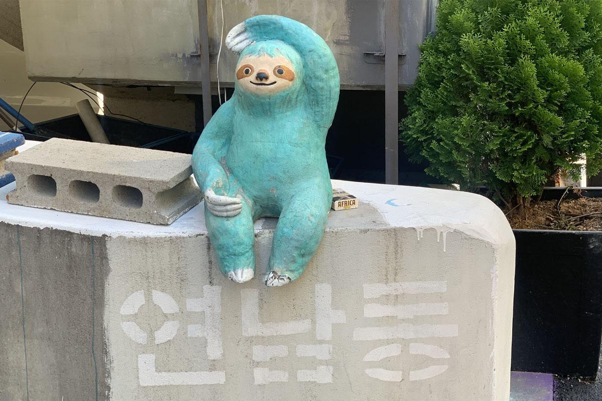 연남동의 잔디밭 옆에 앉아 있는 파란색의 귀여운 나무늘보 조각상과 연남동의 글자가 담긴 콘크리트 벽, 조각상 근처에 놓인 작은 책.