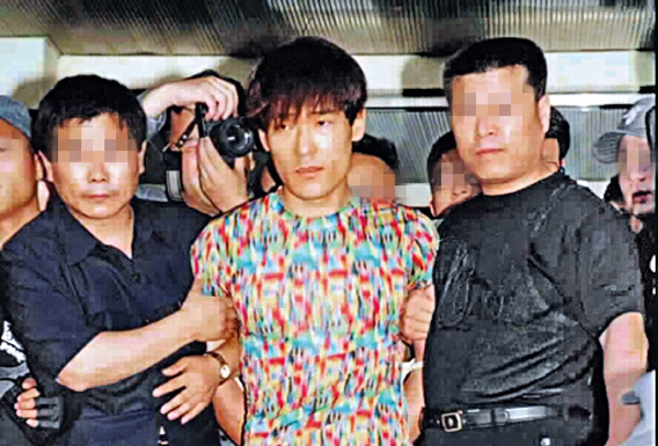 人群中被警察护送的穿彩色图案T恤的角色，场景如新闻取证现场。