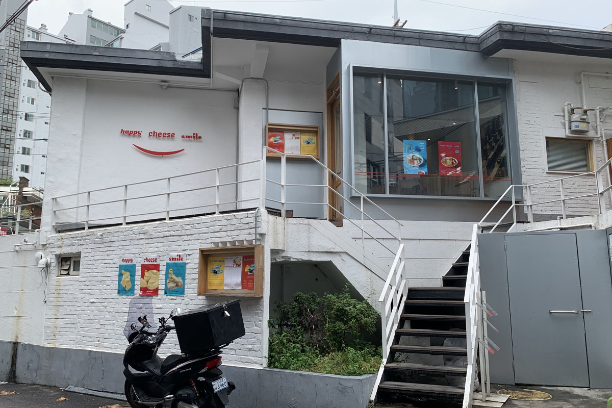 Fachada blanca del restaurante Happy Cheese Smile en Yeonnam-dong, con la entrada al segundo piso visible y carteles coloridos de los menús en las ventanas.