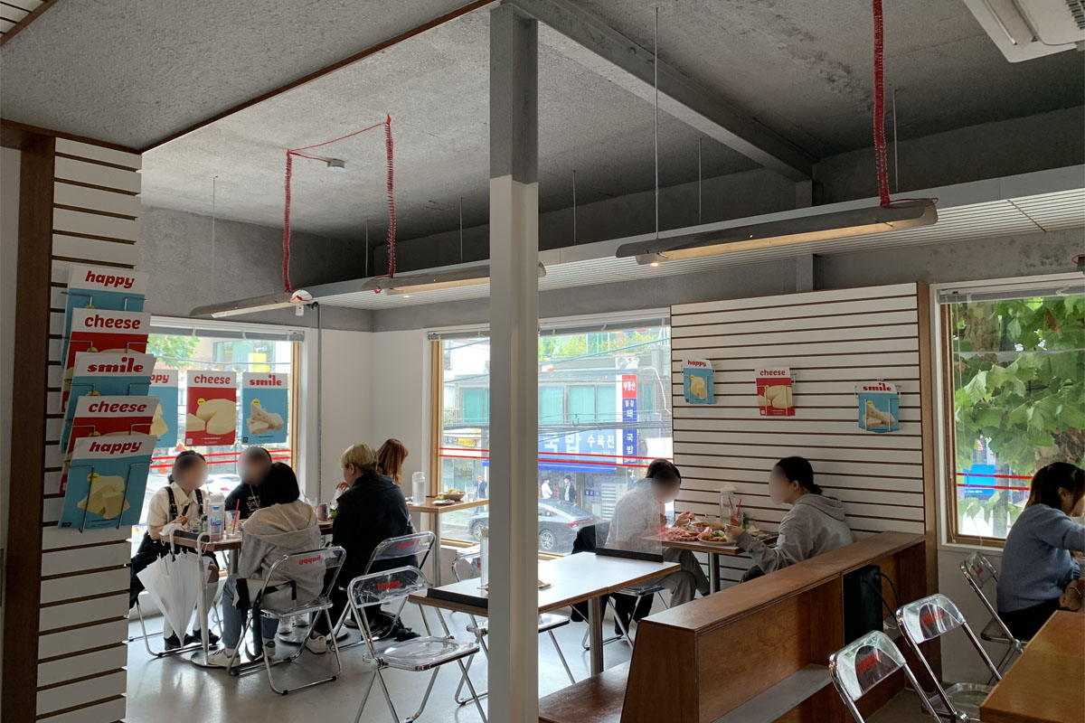 Interior de Happy Cheese Smile, mostrando mesas y sillas modernas con estilo minimalista y personas comiendo, junto a ventanales grandes.