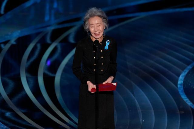 第94回アカデミー賞授賞式でプレゼンターを務めるユン・ヨジョンが手話を交えてスピーチをしている。青いリボンを肩に付け、黒のロングドレスを着用しています。