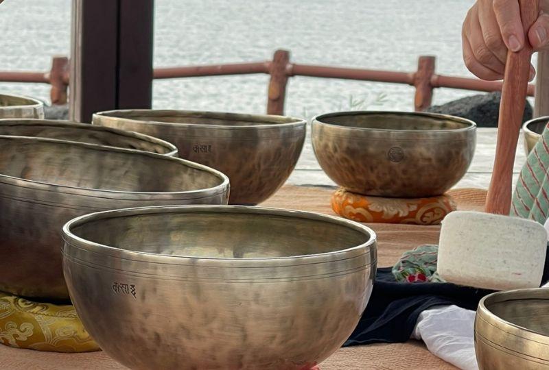 Một người đang cầm một chiếc bát hát Tây Tạng lớn trên bàn trải khăn màu sắc tươi sáng, xung quanh là những chiếc bát khác dùng để trị liệu tinh thần.