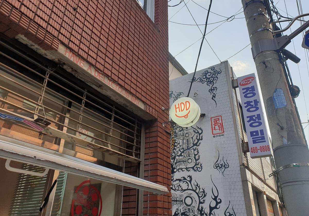 Insegna di HDD Pizza appesa su un muro esterno rosso, con il logo del locale ben visibile accanto a decorazioni artistiche.