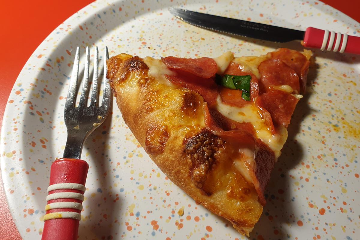 Trancio di pizza al pepperoni su un piatto bianco con posate rosse, evidenziando la texture dorata del bordo.