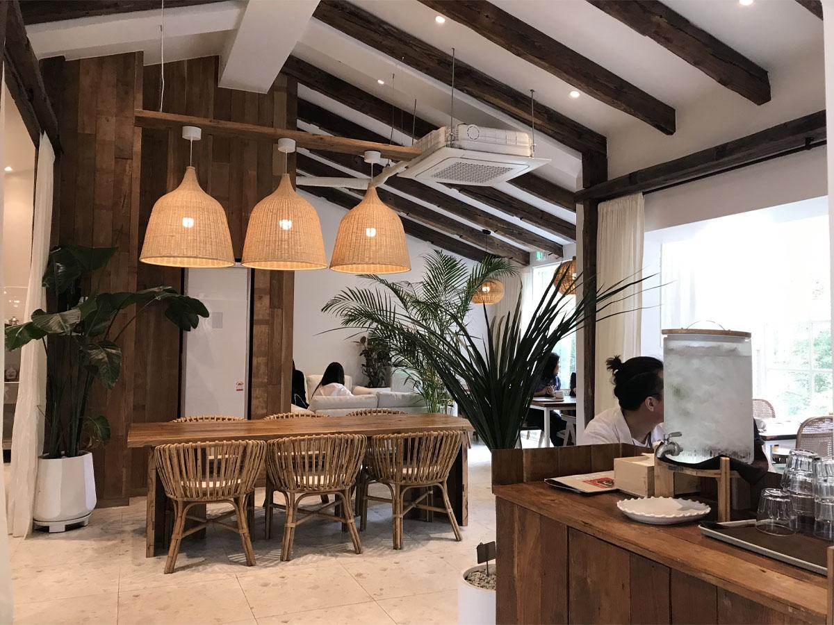 The modern and simple interior of Wake and Bake. Inside you see white walls with wooden accents and pillars accompanied by the rattan furniture and plants.