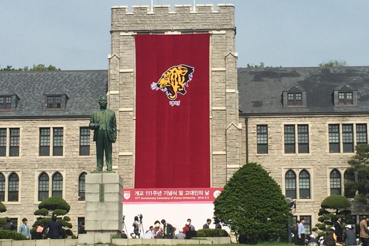 รูปภาพอาคารของมหาวิทยาลัยโคเรียพร้อมด้วยธงรูปเสือบนฉากหลังสีแดงและรูปปั้นบุคคลที่ยืนอยู่ด้านหน้า แสดงถึงสถานที่ทางการศึกษาที่มีชื่อเสียงในประเทศเกาหลีใต้