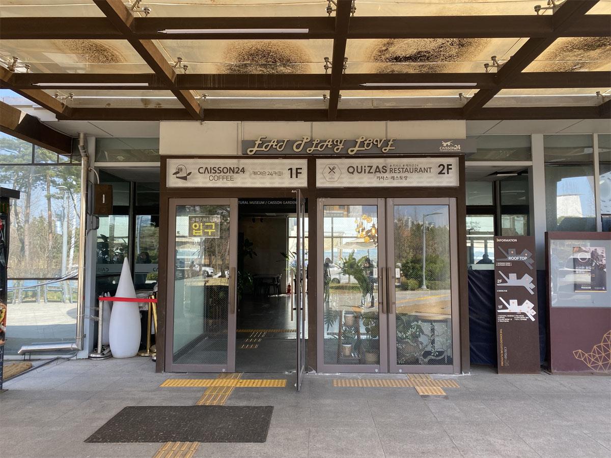 Entrée vitrée du Caisson 24 avec des indications pour le café au premier étage et le restaurant au deuxième étage.