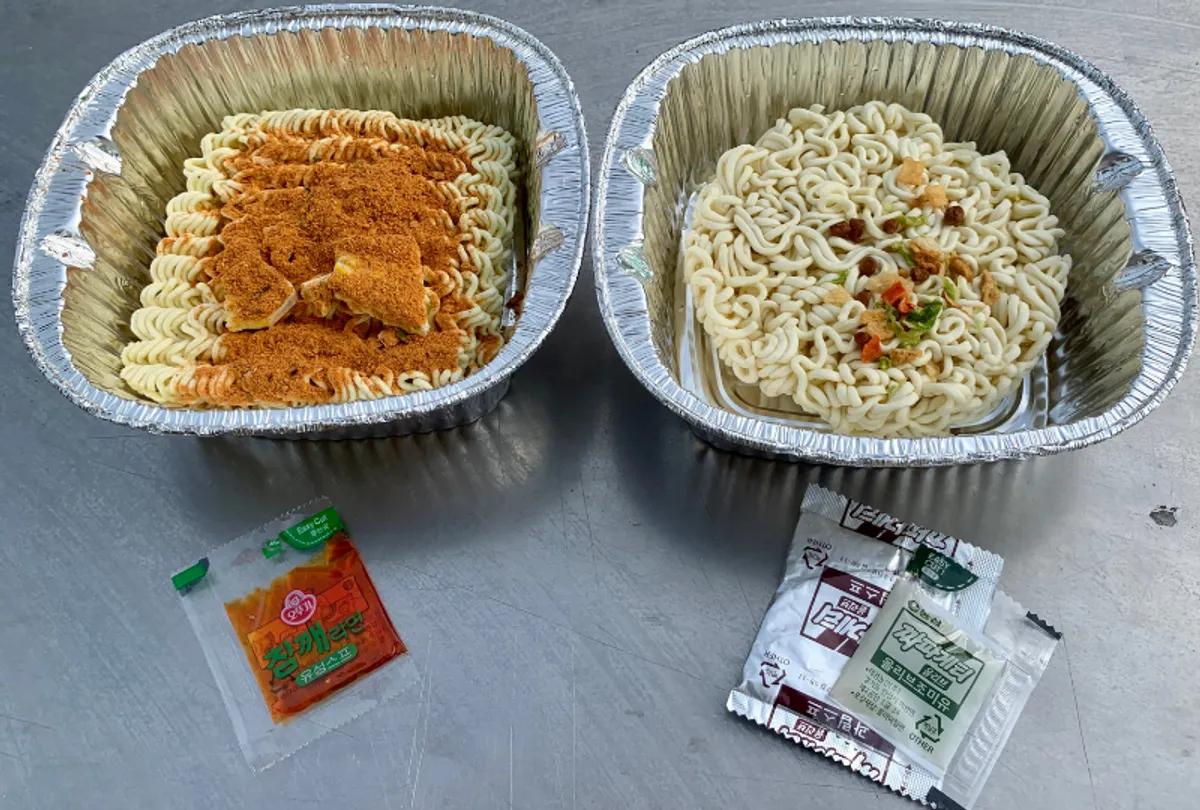 A visual of two bowls of ramen being served, with the cooking process taking place in the background.