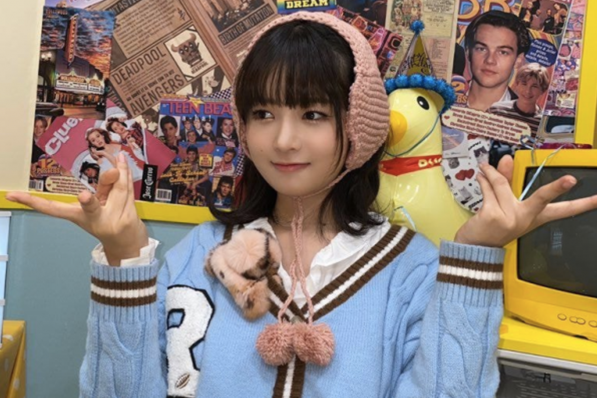 一位穿着蓝色毛衣的女性在复古风墙纸前摆出Gyaru Peace手势,背景布满了怀旧海报和装饰品。