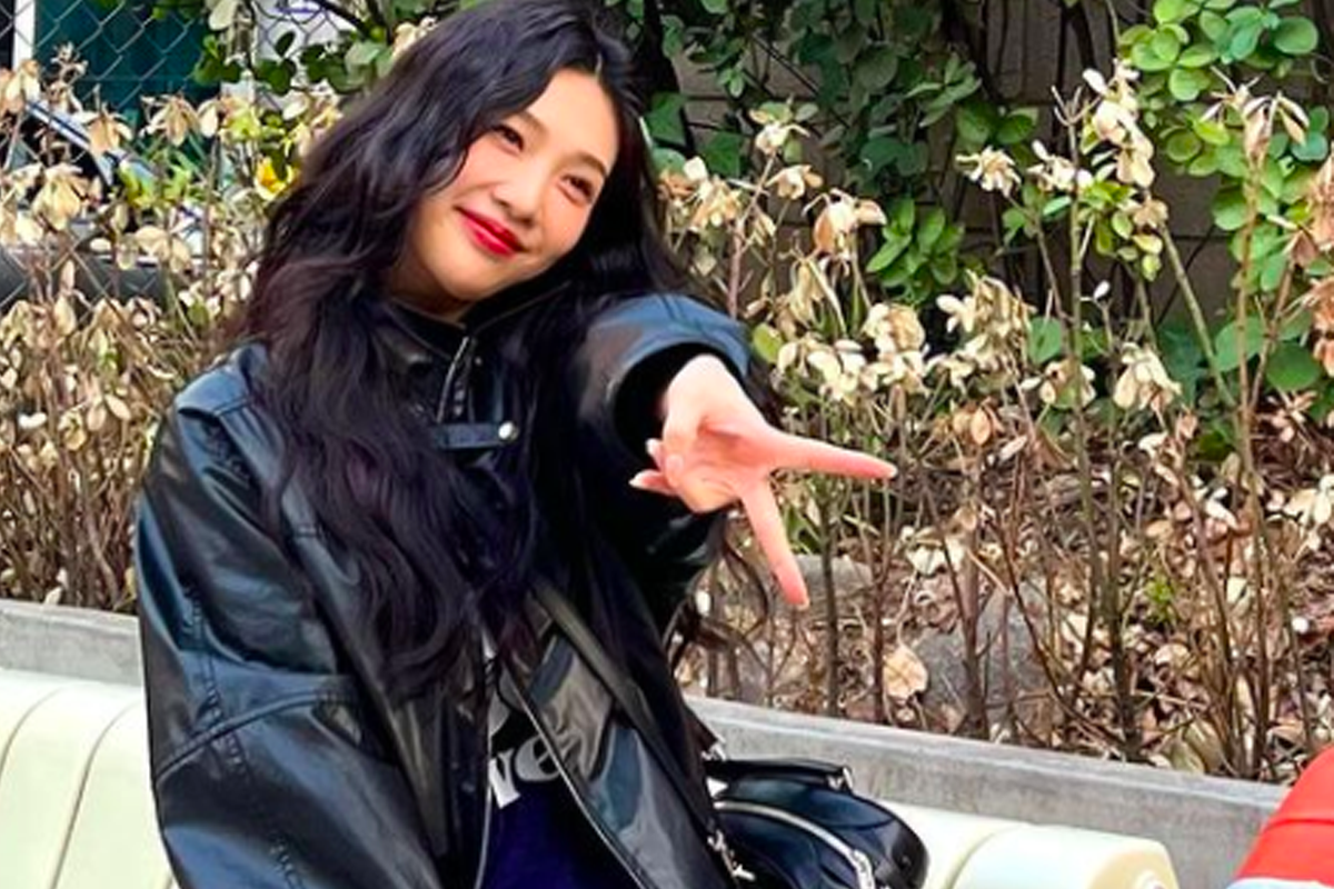 一位穿着黑色皮夹克的女性坐在户外长椅上,展示倒V手势,背景是一片绿树丛。