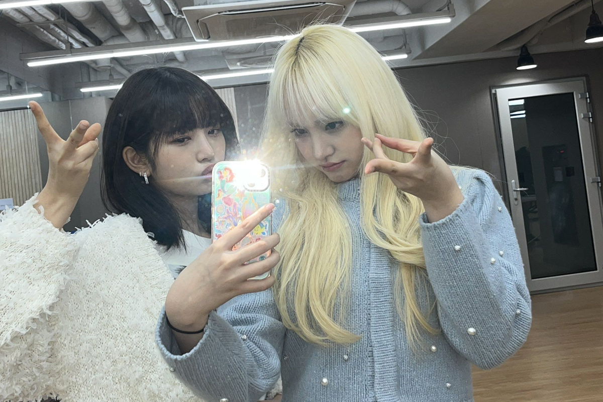 两位韩国艺人在镜子前摆出Gyaru Peace手势,其中一位长发艺人穿着蓝色毛衣,另一位艺人拿着手机自拍。