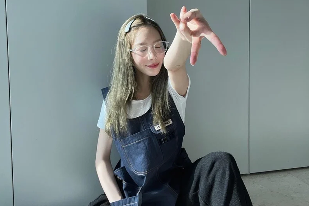 一位穿着牛仔背带裤的长发女性坐在地上伸出手,展现Gyaru Peace手势,背景是现代简约的灰色墙壁。
