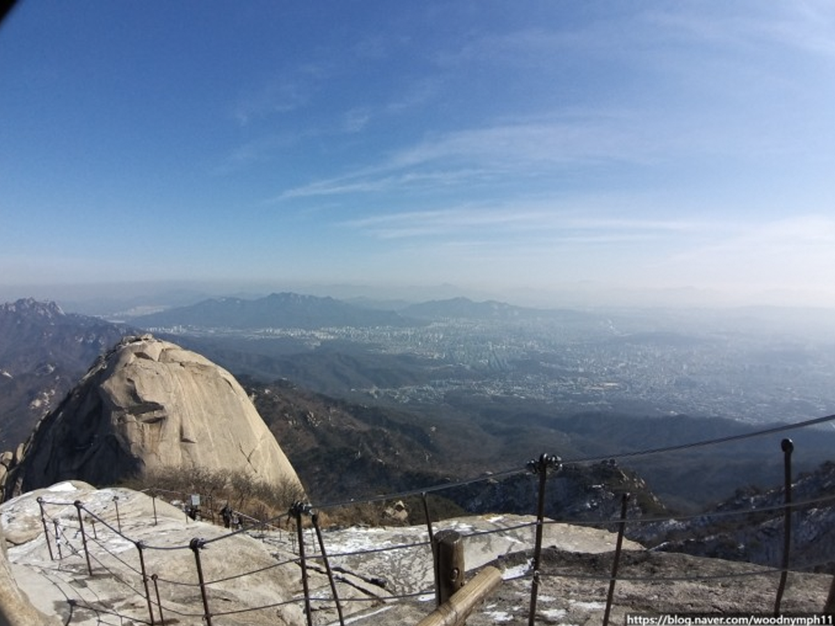 从首尔北汉山顶望向城市的全景，蓝天白云中隐藏着首尔市的建筑群，展现出城市与自然的完美对比。适合SEO优化的全景风光描述。