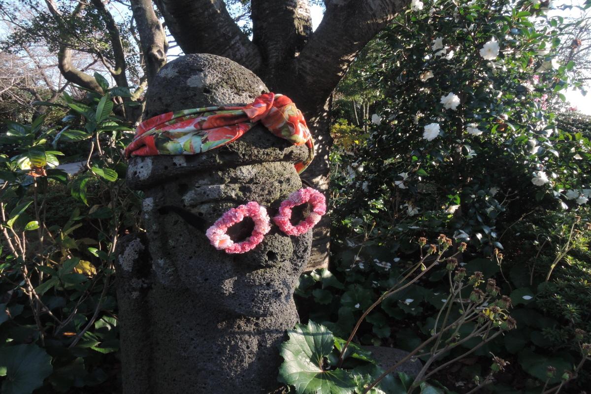 Жежү арлын чулуулаг бэлэг тэмдэг дээр цэцэг бүхий нүдний шил зүүсэн бөгөөд чулууны ард цагаан цэцэгтэй бутнууд харагдаж байна.