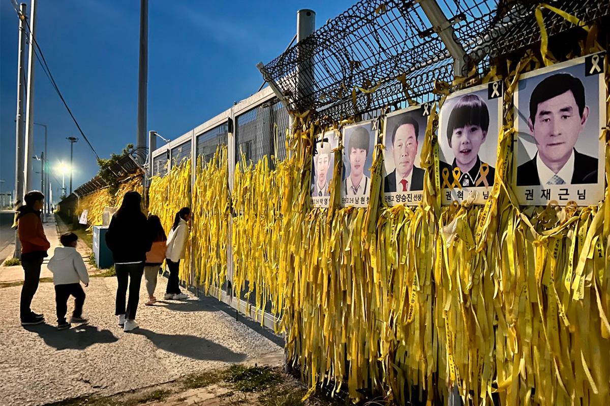 ครอบครัวและผู้เยี่ยมชมยืนรำลึกข้างรูปภาพผู้สูญเสียและริบบิ้นเหลืองในเหตุการณ์เรือเซวอล