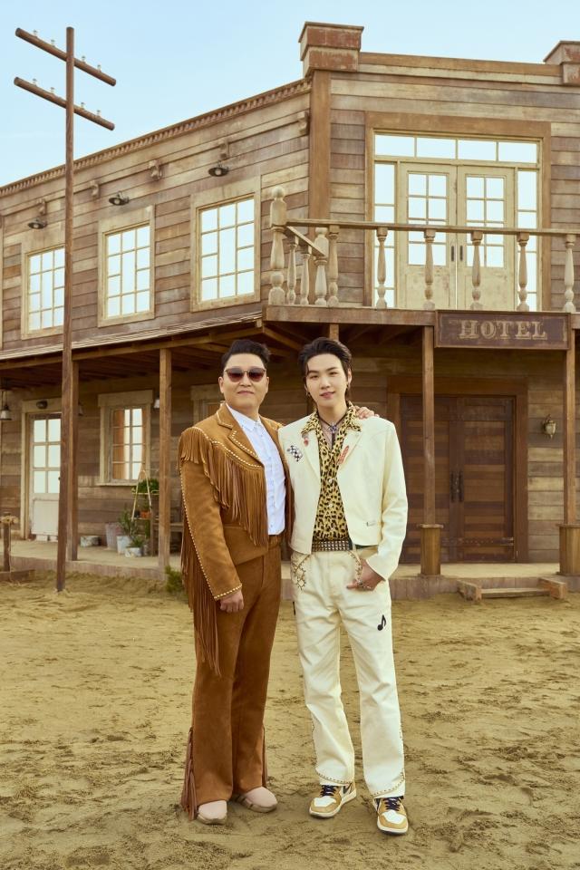 PSYとSUGAが古い西部の衣装を着て楽しそうに写真を撮っている「That That」ミュージックビデオセットの外で立つ姿。木造の建物と砂地の背景が西部スタイルを演出している。