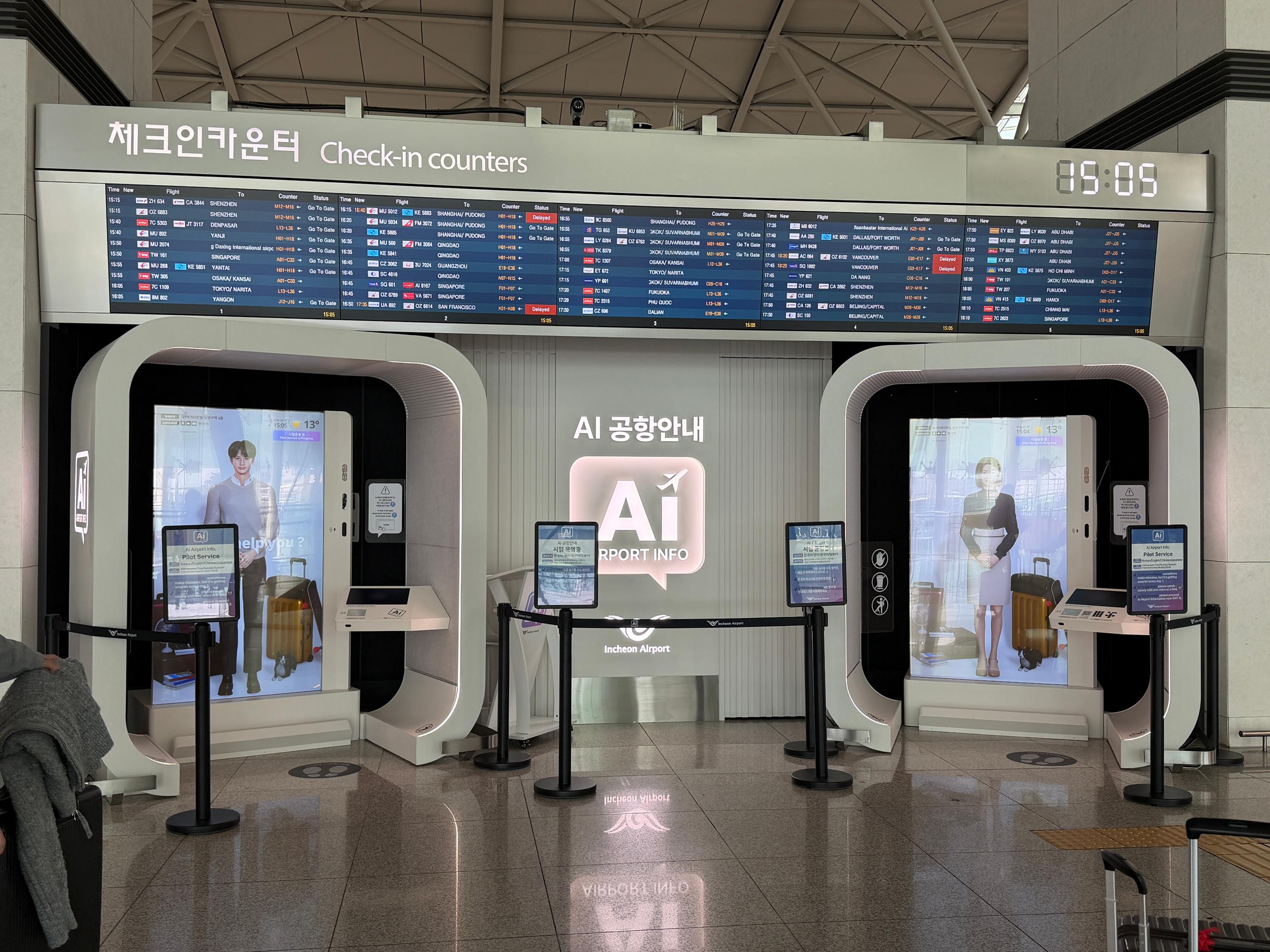 仁川機場第一航廈3樓報到區AI服務站全景，兩個大型互動螢幕展示虛擬客服人形與行李示意，正上方有多列電子航班顯示板，右上角顯示目前時間。場景乾淨明亮，前方設有導引標示與自助服務平板，適合旅客查詢報到與航班資訊。