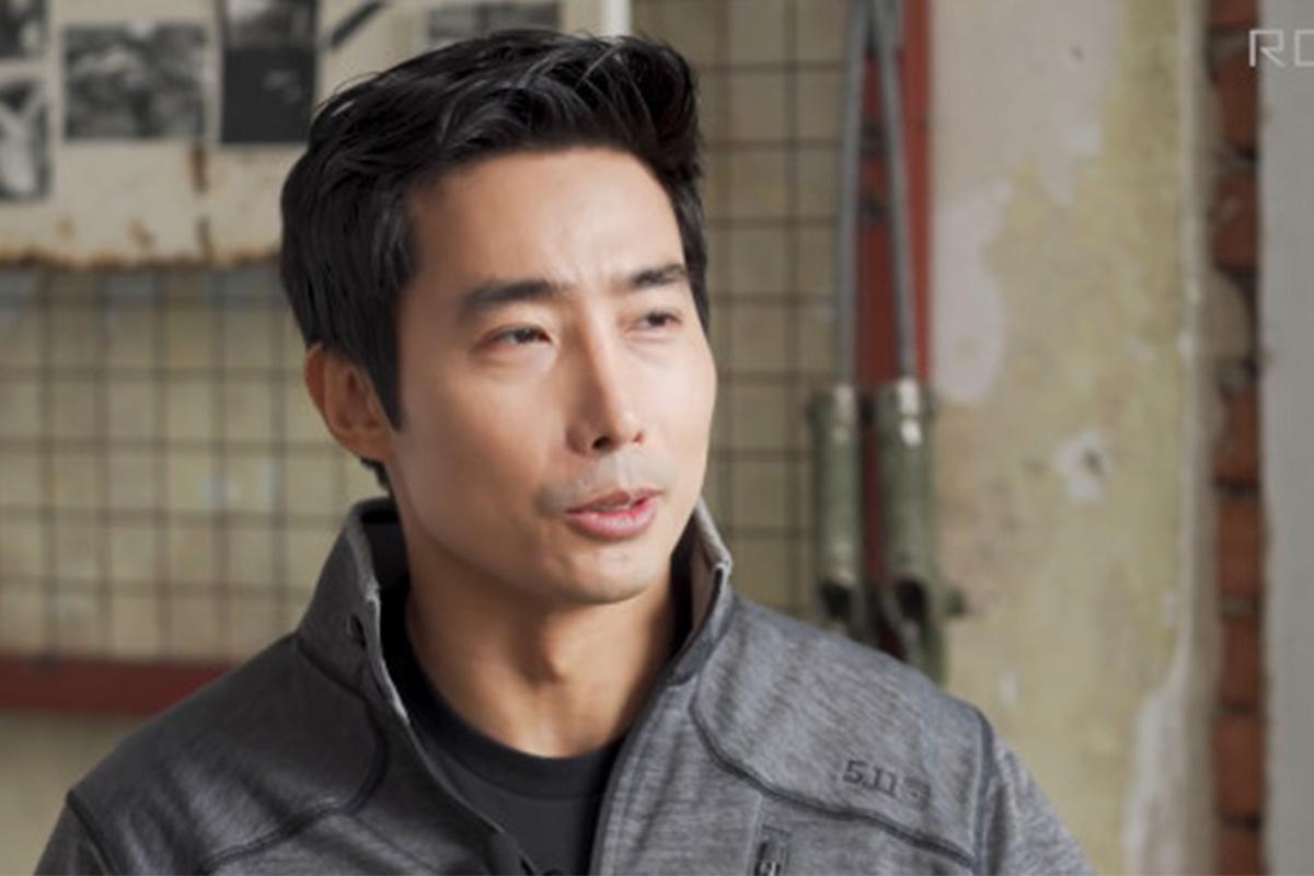 ภาพชายในชุดแจ็กเก็ตสีเข้มอยู่ในสถานที่ที่มีฝาผนังเก่าและกรอบรูปติดอยู่บนกำแพง