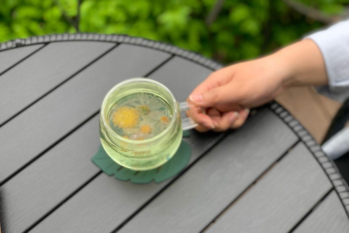 A hand holding a teacup with green tea, set atop a round black table at the Garden of Morning Calm café - emphasizing relaxation amidst nature.