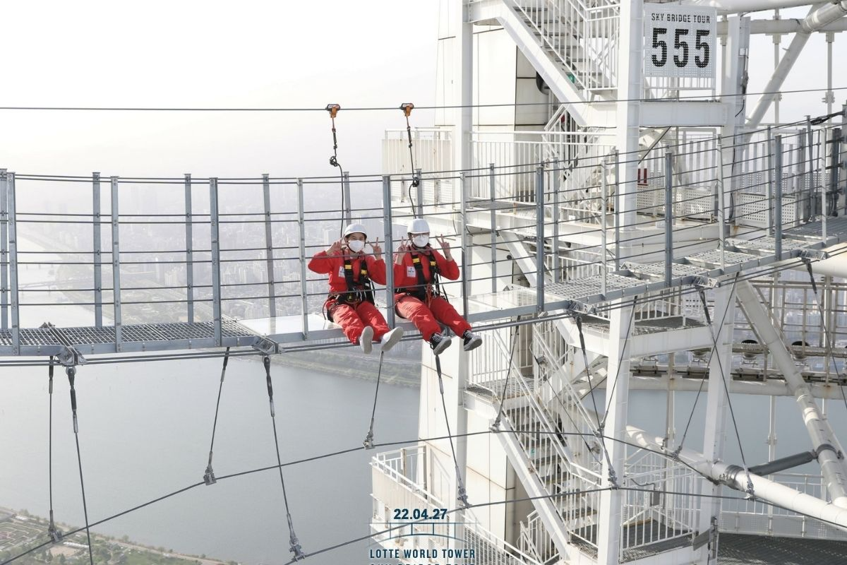 在樂天世界塔頂部的高空行走體驗中，遊客穿著防護服坐在平臺上休息，享受壯觀的城市全景，提升旅遊體驗的獨特性。
