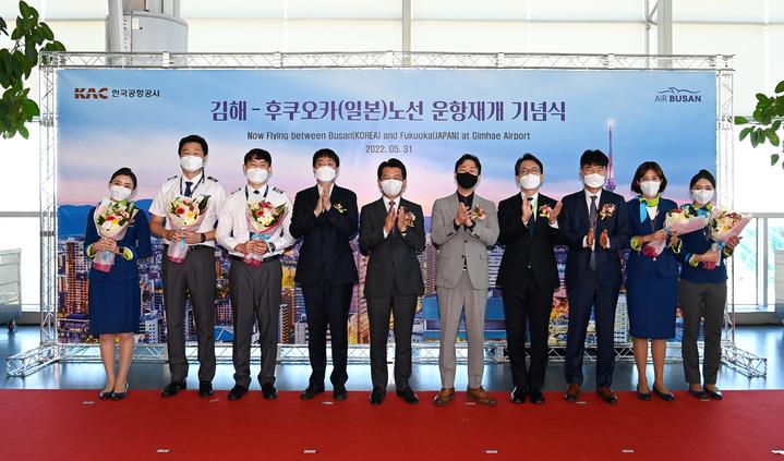 金海空港で行われた金海〜福岡ルート運航再開記念式で、出席者が壇上に並んで記念写真を撮影している様子。背景には釜山の風景が描かれた大きな看板が設置されている。