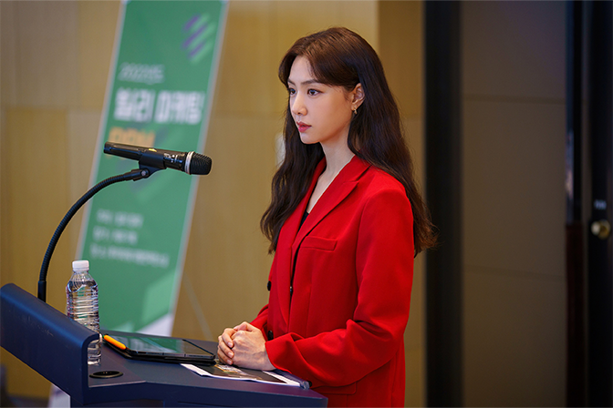 韩剧《第六感之吻》中，角色身穿红色套装正在发表演讲，显示一种职场职能。背后有一块绿色背景板，上面显示着会议的相关信息。