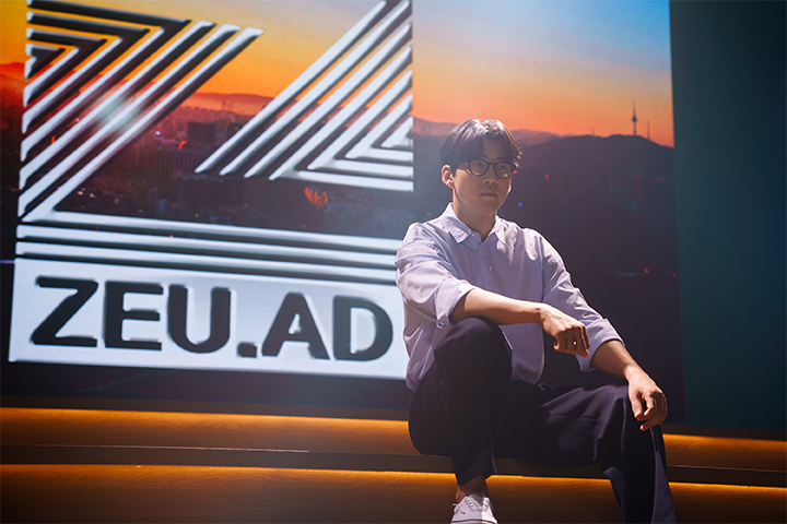 《第六感之吻》的剧照，一位角色坐在台阶上，背景是一个显示ZEU.AD标志的大屏幕，城市天际线和日落的画面。