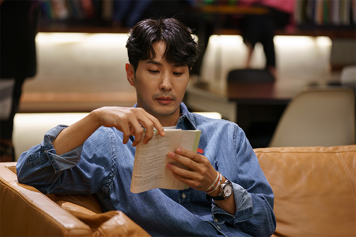 电视连续剧《第六感之吻》中，一位角色在阅读一本书，穿着休闲牛仔衬衫，坐在舒适的沙发上，营造出一种轻松的家庭环境。