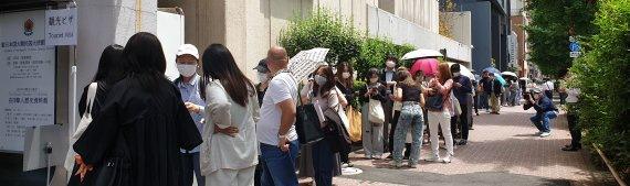 東京の韓国領事館前で観光ビザを申請するため並ぶ人々の列。初夏の晴天の下、日傘や帽子を使いながら、若者や中年の方まで様々な年齢の人々が見られる。