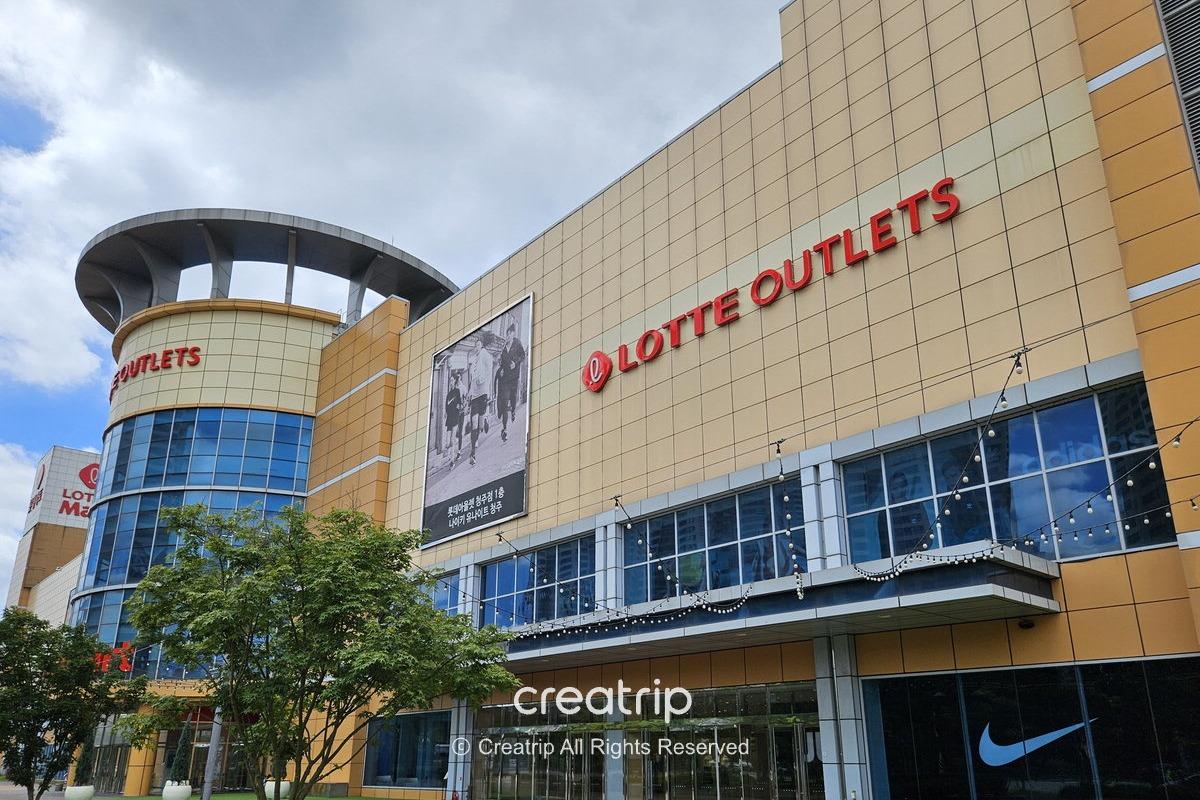韓國大型購物中心外觀：黃色方格外牆與玻璃窗，外牆紅色「LOTTE OUTLETS」招牌，門口樹木與晴天雲景，示意Outlet可能營業或公休日前查詢營業資訊。