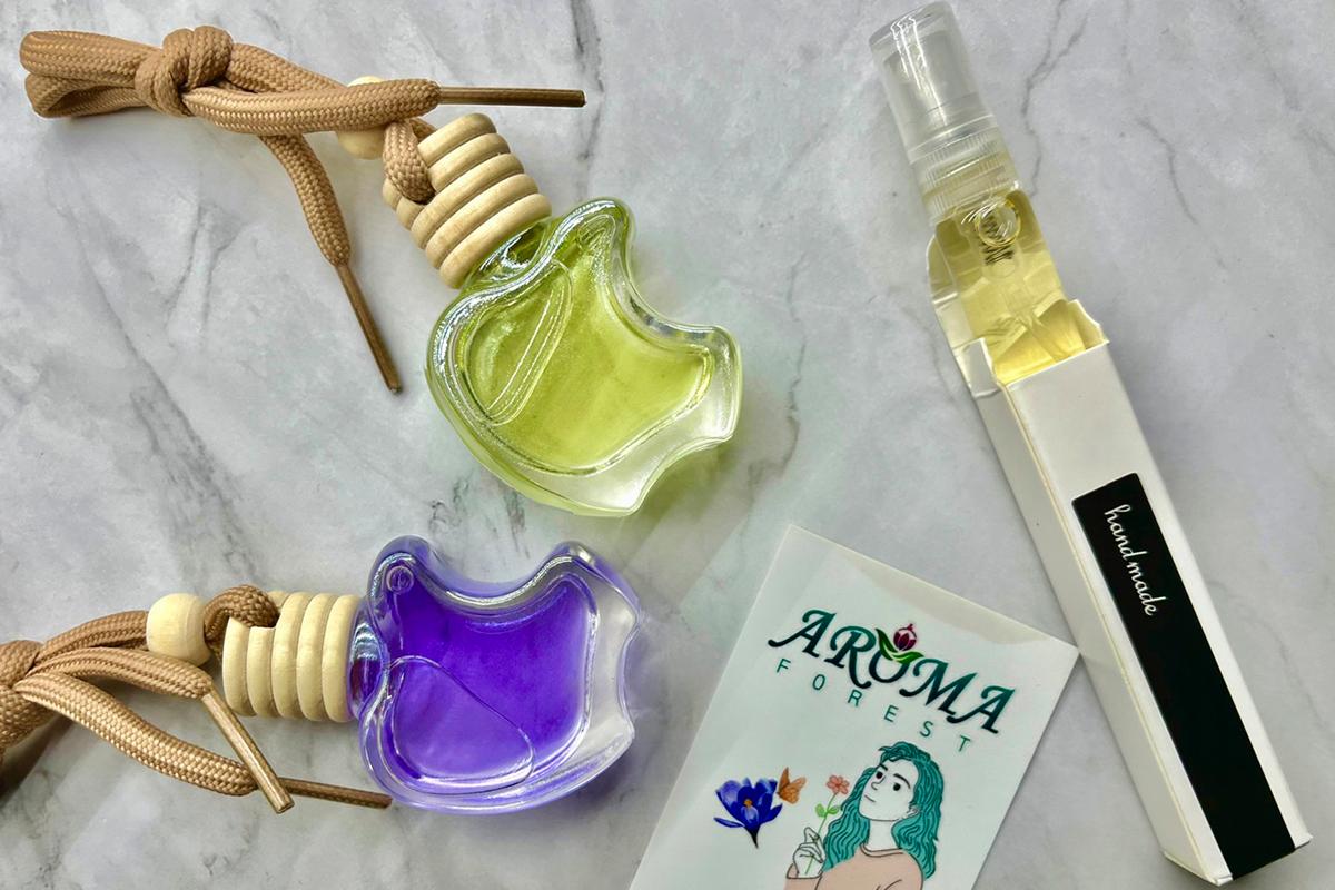 ขวดน้ำหอมสองขวดสีเขียวและสีม่วงจาก Aroma Forest พร้อมบัตรและขวดน้ำมันหอมระเหยสำหรับรถยนต์ วางบนพื้นหินอ่อน
