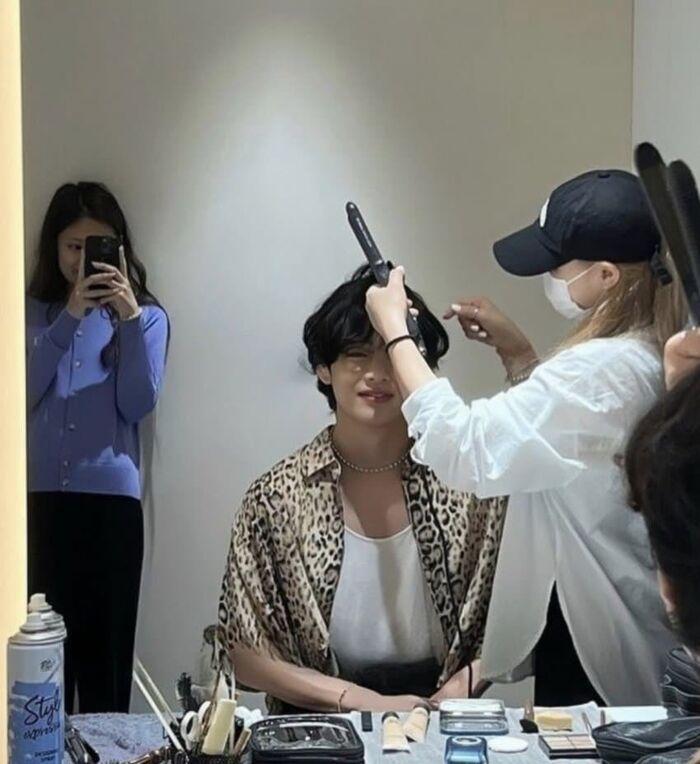 鏡の前の男性が美容師からヘアスタイリングを受けている。女性が後ろからその様子をスマートフォンで撮影しようとしている。男性はヒョウ柄のシャツを着ている。