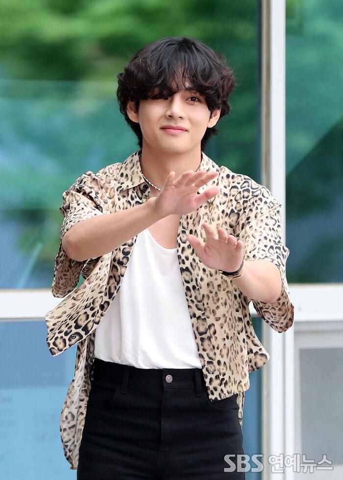 ヒョウ柄のシャツを着た男性が屋外で手を挙げている。背景には緑の自然が見える。