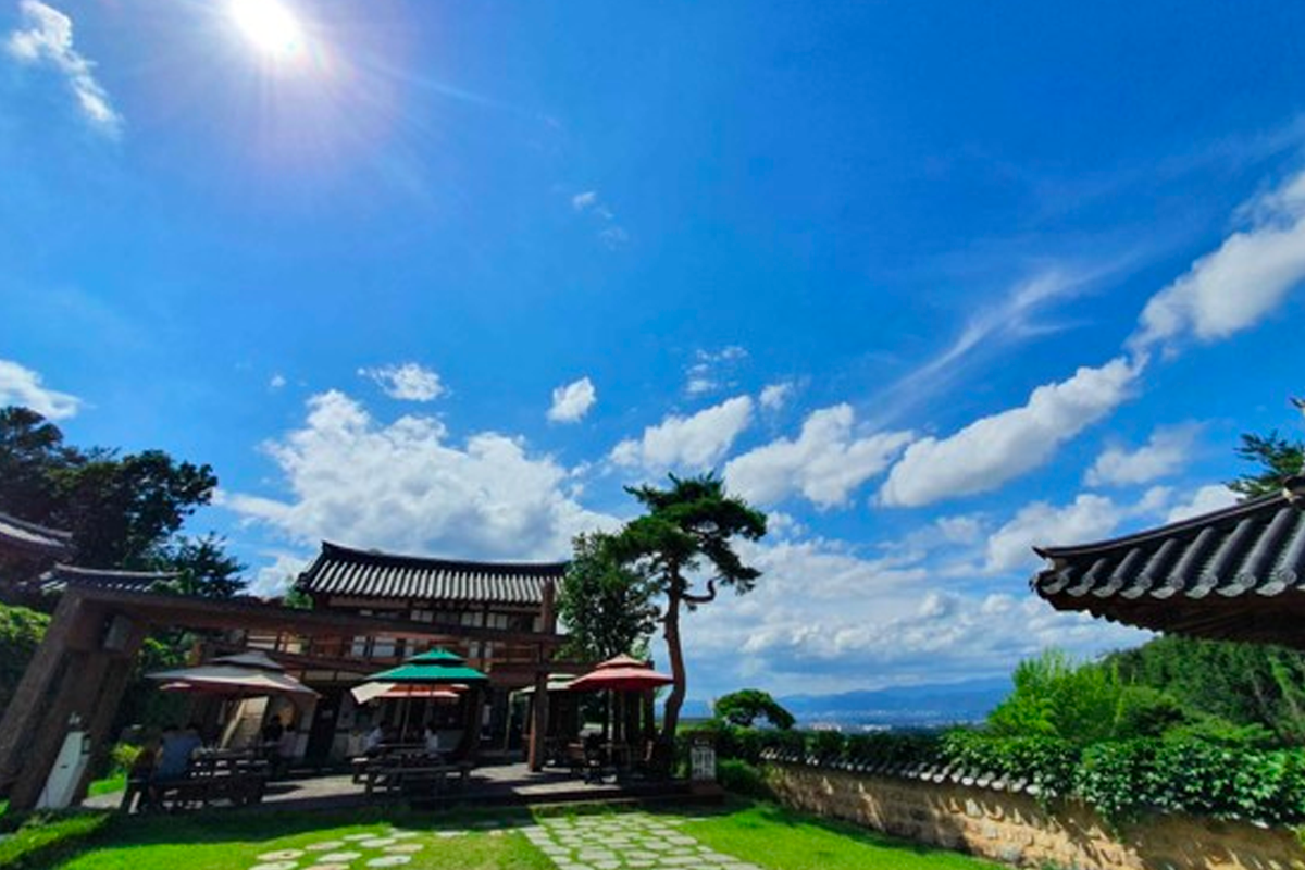 阳光明媚下的韩屋前庭，木桌椅整齐排列，供游客休息，享受山林景色。