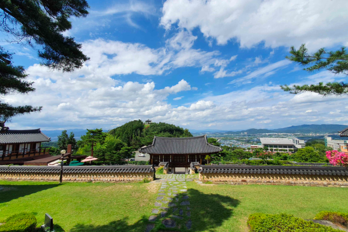 传统韩屋坐落在绿意盎然的山丘上，蓝天白云相伴，远处可见山景。