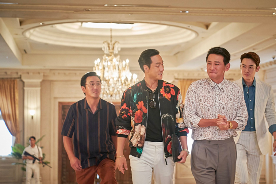 一群男性邁步走在豪華的室內空間中，穿著講究且各自擁有鮮明的風格，華麗的燈飾增加了場景的印象。這是《毒梟聖徒》中的一個場面。