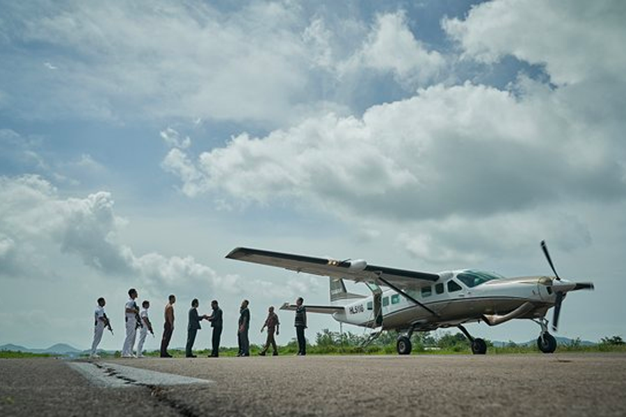 一群人在機場跑道上走向停留的飛機，背景是蔚藍的天空，場景營造出即將展開行動或逃離的氣氛。《毒梟聖徒》中的飛機場景。