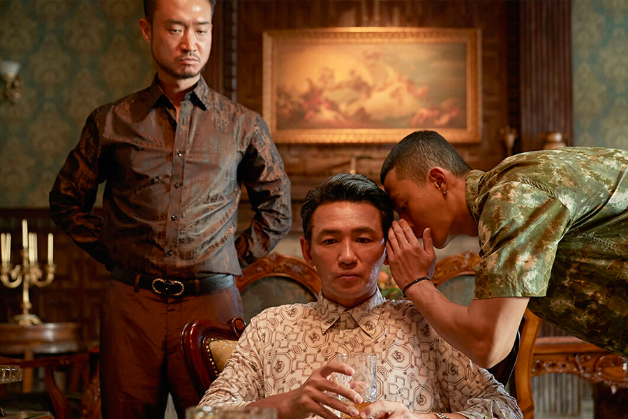 在華麗的室內裝潢中，幾位穿著正式的男子圍繞在一個被蒙面的人旁邊，場面顯示出一種緊張且重要的情節。《毒梟聖徒》中的關鍵對話場景。