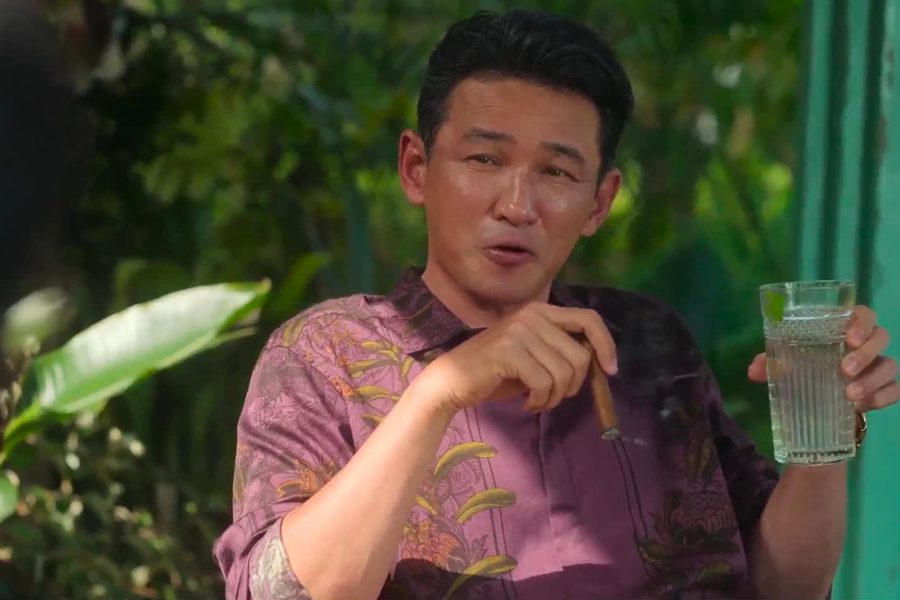 一名身穿色彩豐富的襯衫，手持一杯水和雪茄的男子，坐在戶外，背景則是一片綠色植栽，營造出悠閒舒適的氛圍。《毒梟聖徒》劇照之一。