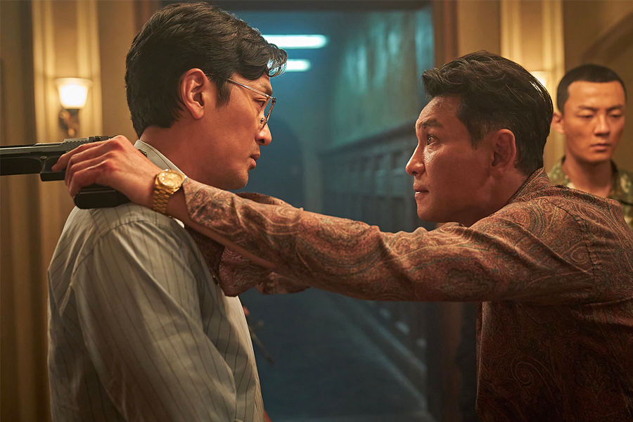 一名男子手持手槍指向另一名男子，背景是幽暗的走廊，這場景充滿了緊張和驚險感。《毒梟聖徒》中的緊張對峙。