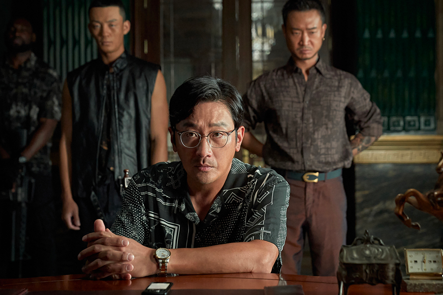 一位穿著印花襯衫的男子坐在桌前，雙手交疊，背景有兩位穿著深色服裝的男子，創造出一種權力和神秘的對峙感。《毒梟聖徒》中的場景。