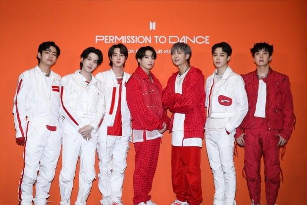 BTSメンバーがオレンジ色の背景の前に立っている。彼らは白と赤の衣装を着ており、グループのパフォーマンス衣装を身に纏っている。