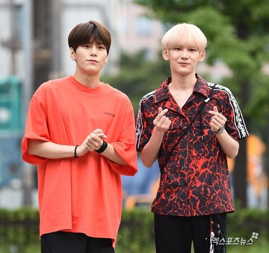 韓国男性アイドルグループJBJ95のメンバー、明るい色の衣装を着て指を使いハートを作るジェスチャー。背景は街の景色。