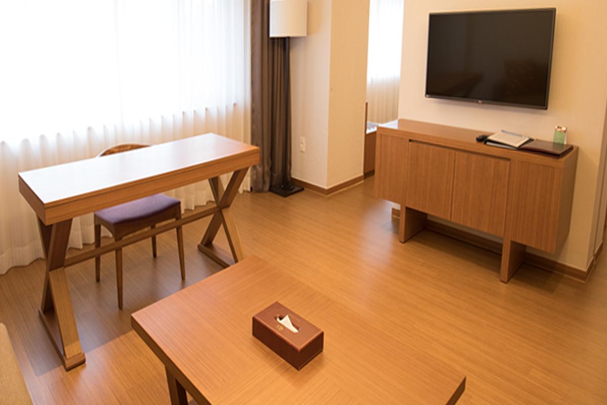 Living area in a hotel suite with a work desk, TV, and wooden flooring.