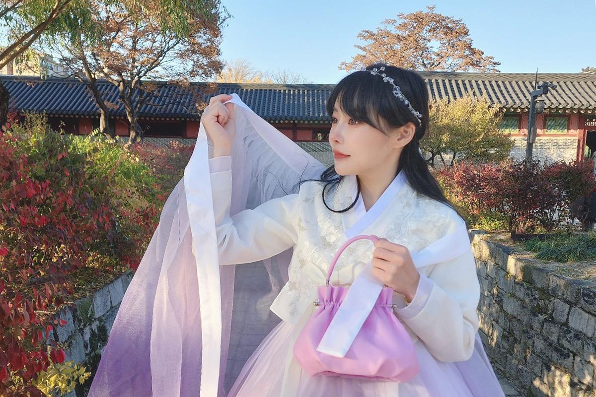 전통 한복을 입은 여성이 서울의 전통 궁궐 앞에서 포즈를 취하고 있는 모습. 흰색 저고리와 보라색 치마가 어우러져 우아하고 매혹적입니다.