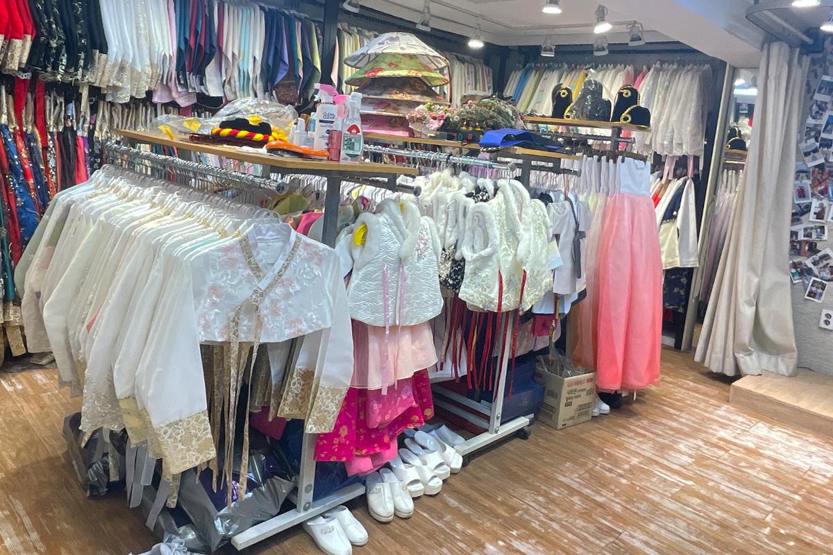 서울의 한복 대여점 내부 전경. 다양한 색상과 스타일의 한복들이 여러 개의 옷걸이에 걸려 있으며, 선반에는 전통 모자와 장식품들이 놓여 있습니다.