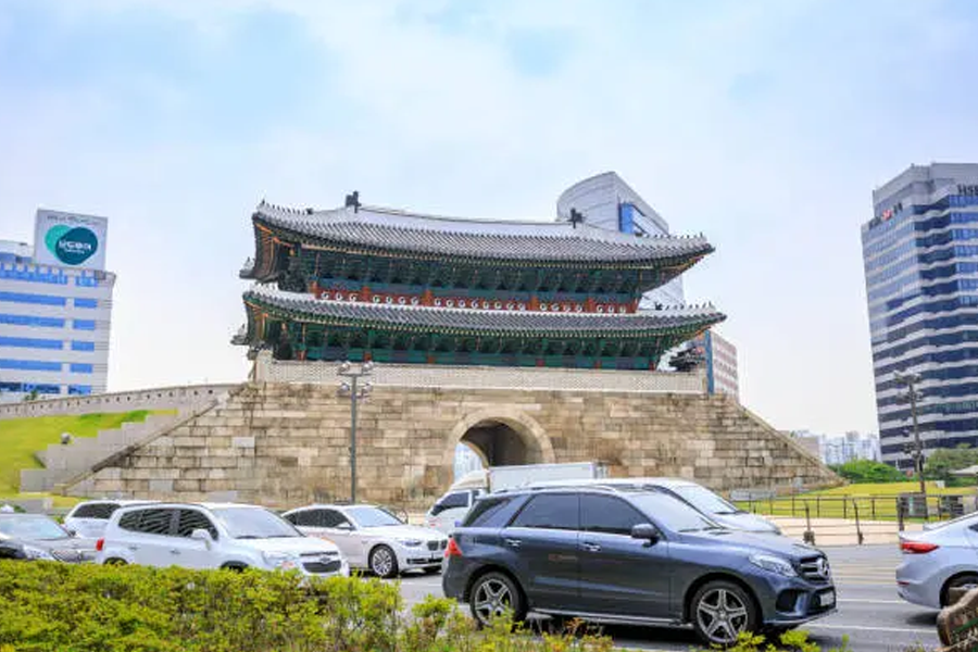 한국 전통 건축 양식이 적용된 건물 앞에 다양한 차량이 주차되어 있는 역동적인 서울 시내 모습입니다. 서울 여행 및 관람차 서비스를 홍보하는데 쓸 수 있습니다.