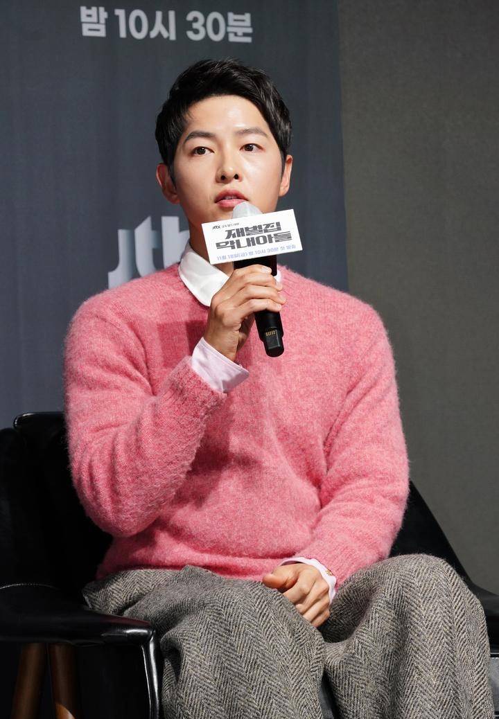 ソン・ジュンギ、ピンクセーターと白シャツを着用し、インタビュー中の様子。韓国の俳優として知られ、最近の交際報道で注目されている。抱いているマイクには『財閥家の末息子』と書かれている。