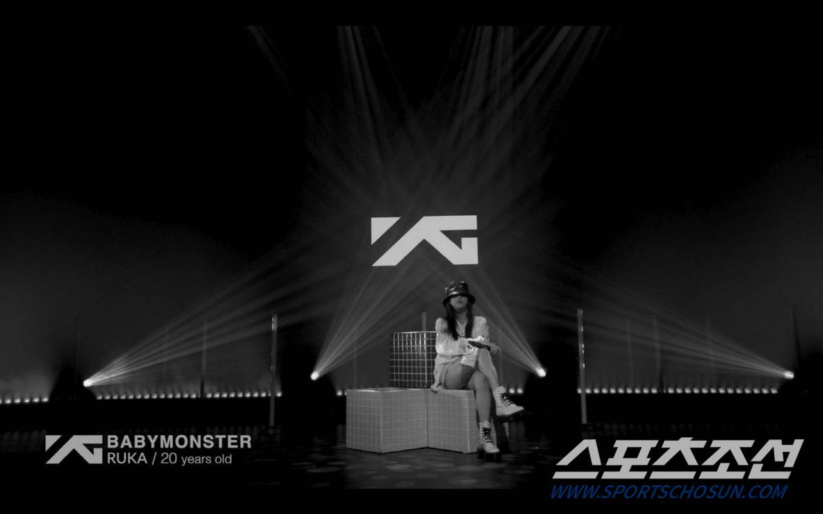 新しいガールズグループBABYMONSTERのルカ、YGのロゴを背景に座っている。